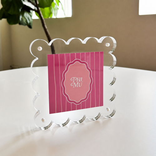 The Darling Effect's Phi Mu Square Scalloped Acrylic Frame displays a pink PHI MU card in white script on a table with a leafy plant and window, adding charm to your modern picture frames collection.
