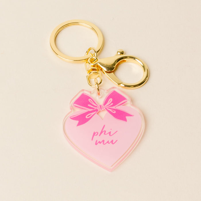 The Darling Effect's Phi Mu Sweetheart Bow Acrylic Keychain features a pink heart with "phi mu" in pink script, a pastel bow design, and a gold keyring—perfect as a sorority gift.