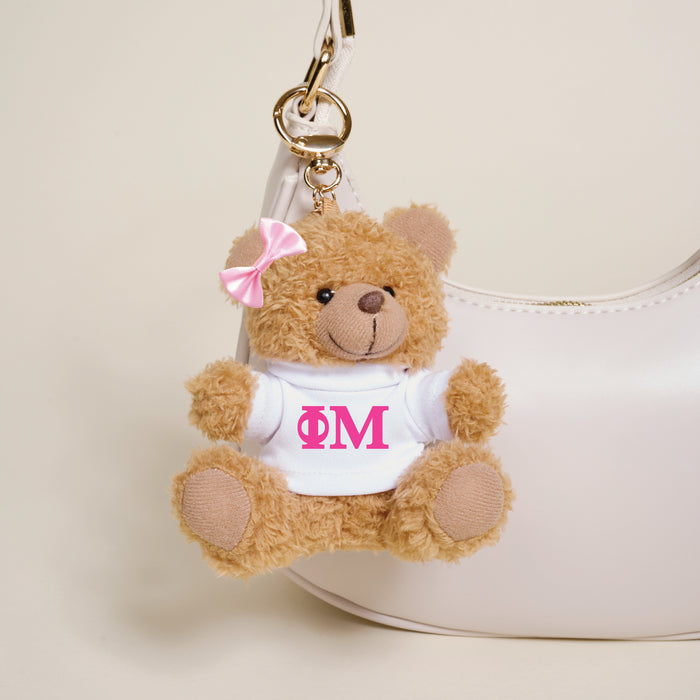 The Darling Effect’s Phi Mu Teddy Bear Bag Charm—a small brown bear with a pink bow and Greek letters on its white shirt—hangs from a white handbag, making it a charming sorority accessory or thoughtful gift.