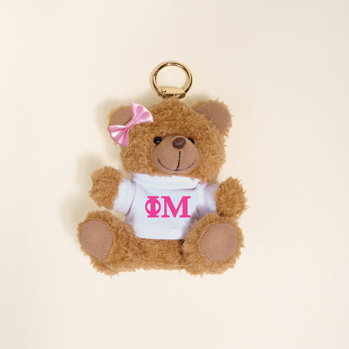 The Darling Effect’s Phi Mu Teddy Bear Bag Charm is a small brown teddy bear keychain with a pink bow and white shirt featuring pink ΦΜ letters, making it a cute sorority gift. Photographed on a light beige background.