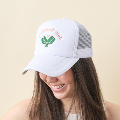 A smiling woman with long brown hair wears the-darling-effect’s Pickleball Club Trucker Hat, a white mesh cap with embroidered lettering, two green pickleball paddles, a small heart, and “Pickleball Club” on the front.