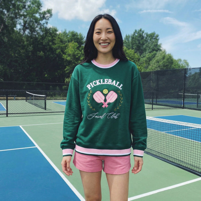 A smiling woman stands on a pickleball court wearing The Darling Effect’s Pickleball Social Club Oversized Crew, featuring embroidered pickleball designs—a cozy, stylish staple that pairs perfectly with her pink shorts.