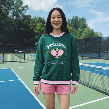 A smiling woman stands on a pickleball court wearing The Darling Effect’s Pickleball Social Club Oversized Crew, featuring embroidered pickleball designs—a cozy, stylish staple that pairs perfectly with her pink shorts.
