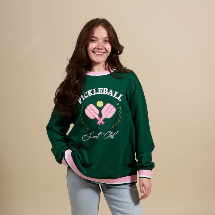 A young woman with long brown hair smiles, wearing The Darling Effect’s Pickleball Social Club Oversized Crew in green with pink trim, featuring embroidered pickleball and "Pickleball Social Club" details.