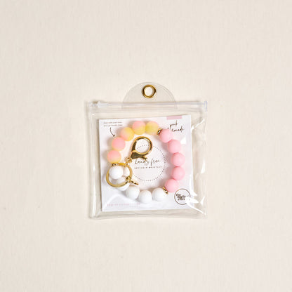 The Darling Effect’s Pink Lemonade Hands-Free Keychain Wristlet features pink, yellow, and white silicone beads with a gold keyring and clasp, shown in a clear pouch on a light beige background for stylish convenience.