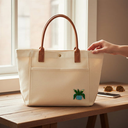 A cream-colored canvas tote from The Darling Effect sits on a table, featuring the Plant Mix & Match Iron-On Patch—a small green and blue embroidered plant—perfect for customizing with more patches or designs.