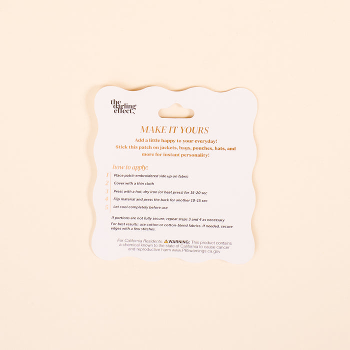 A square, scalloped cream card from The Darling Effect displays instructions for the Plant Mix & Match Iron-On Patch with orange and black text detailing steps, warnings, and a website link for customizing items with colorful designs.