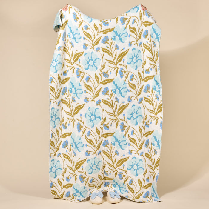 A person stands wrapped in The Darling Effect's Poppin' Petals Aqua Cozy Dreams Luxe Blanket, featuring blue and green floral designs, with only their hands and white shoes showing against a beige background.