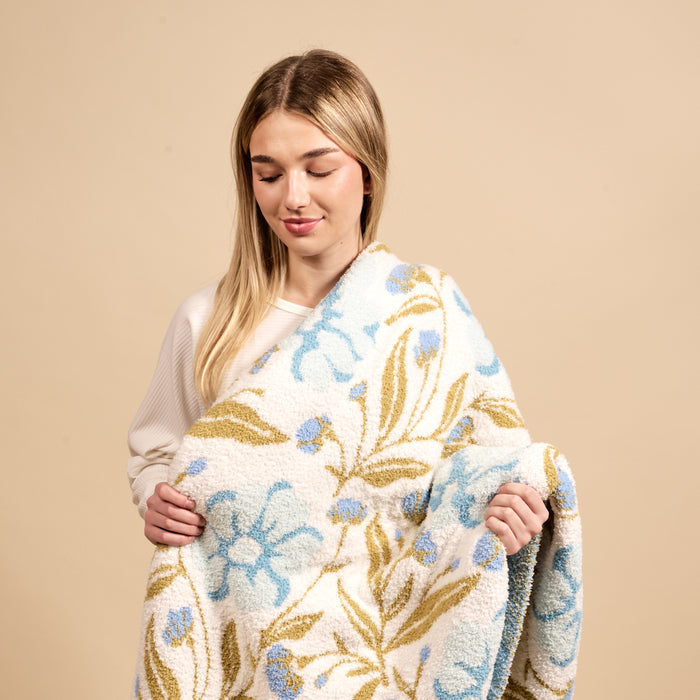 A smiling young woman with blonde hair wraps herself in The Darling Effect’s Poppin' Petals Aqua Cozy Dreams Luxe Blanket, an ultra-soft throw featuring a blue floral and olive green leaf pattern on a neutral beige background.