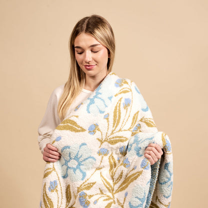 A smiling young woman with blonde hair wraps herself in The Darling Effect’s Poppin' Petals Aqua Cozy Dreams Luxe Blanket, an ultra-soft throw featuring a blue floral and olive green leaf pattern on a neutral beige background.