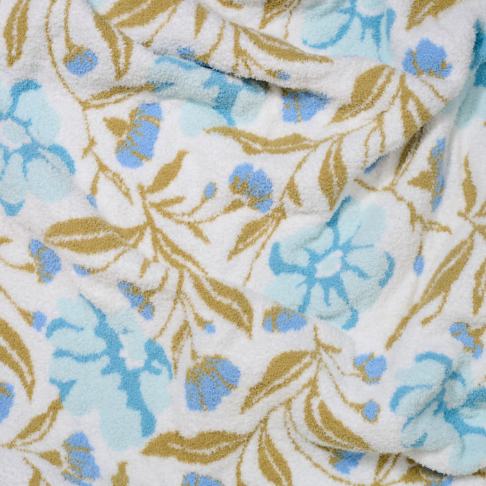 A close-up of the ultra-soft Poppin' Petals Aqua Cozy Dreams Luxe Blanket by The Darling Effect, showing its textured fabric with a floral pattern of light blue flowers and tan leaves on a white background, gently folded.