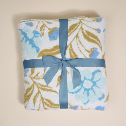 A folded Poppin' Petals Aqua Cozy Dreams Luxe Blanket by The Darling Effect, featuring blue and gold florals and tied with a blue ribbon, is displayed on a light beige background—an ultra-soft touch of elegance for any space.