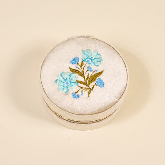 The Darling Effect's Poppin' Petals Aqua Round Velvet Jewelry Case features embroidered blue flowers and green stems on the cream-colored zippered lid, shown against a plain light beige background.