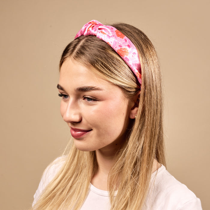 A young woman with long blonde hair smiles slightly and looks to the side, wearing The Darling Effect's Poppin' Petals Hot Pink Classic Twist Headband and a white shirt against a plain beige background.