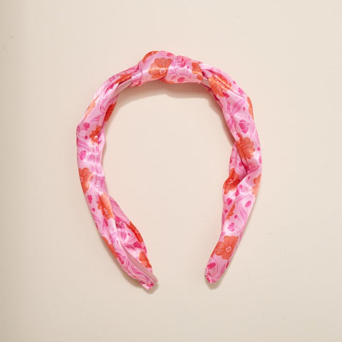 The Darling Effect's Poppin' Petals Hot Pink Classic Twist Headband, featuring bold orange and red floral patterns on pink fabric, is displayed on an off-white background—a stylish accessory for any occasion.