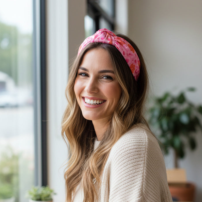 A woman with long wavy hair wears The Darling Effect Poppin' Petals Hot Pink Classic Twist Headband and a cream sweater, smiling by a window with indoor plants in the background.