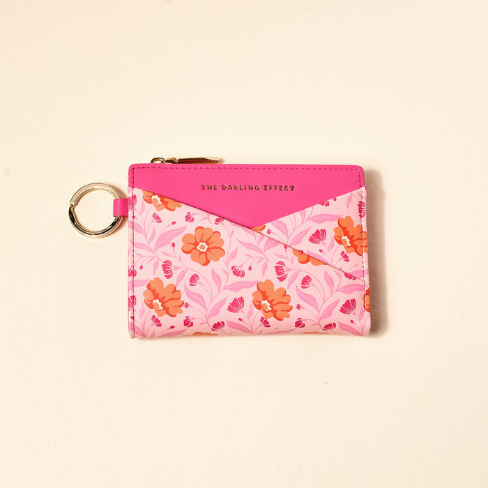 The Darling Effect Poppin' Petals Hot Pink Luxe Pocket Wallet features an orange and pink floral pattern, a key ring, gold zipper, and "THE DARLING EFFECT" printed on a solid pink flap—an essential for everyday use.