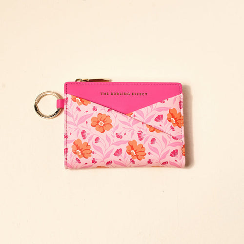 The Darling Effect Poppin' Petals Hot Pink Luxe Pocket Wallet features an orange and pink floral pattern, a key ring, gold zipper, and "THE DARLING EFFECT" printed on a solid pink flap—an essential for everyday use.