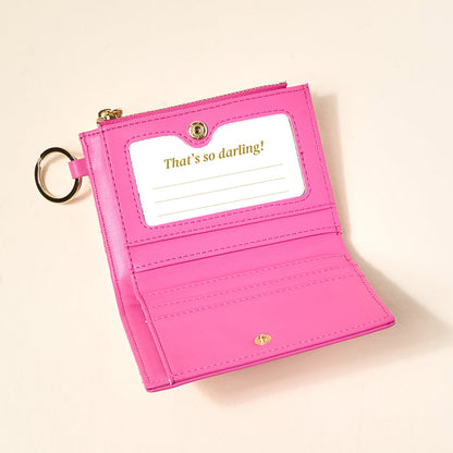 The Darling Effect's Poppin' Petals Hot Pink Luxe Pocket Wallet, crafted from vegan leather, features a keyring, card slots, and a clear ID window with a gold "That's so darling!" card—an everyday essential on a light beige surface.