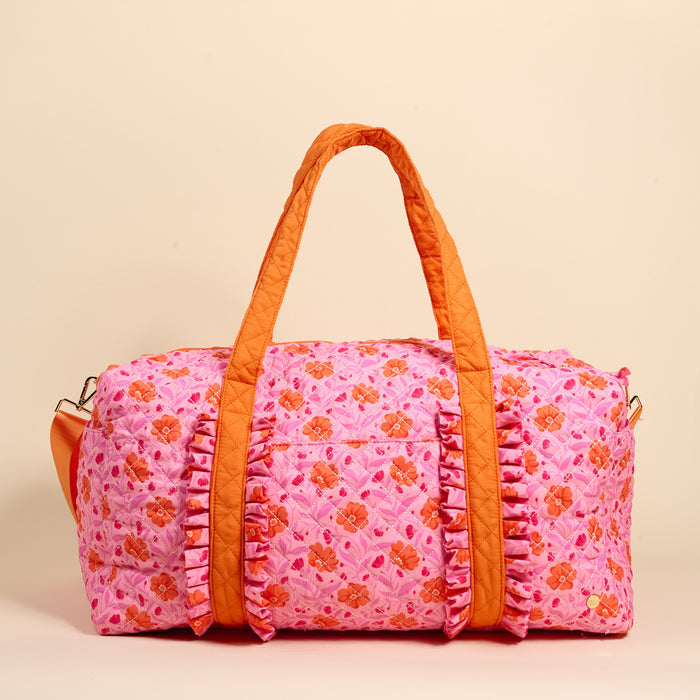 The Darling Effect's Poppin' Petals Hot Pink On-the-Go Quilted Weekender Bag features an orange flower pattern, matching handles, and ruffled trim—ideal as a spacious, stylish travel bag for your next adventure.