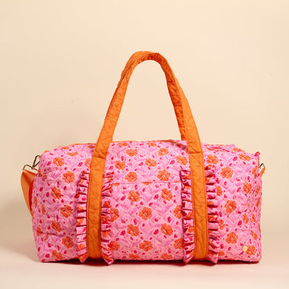 The Darling Effect's Poppin' Petals Hot Pink On-the-Go Quilted Weekender Bag features an orange flower pattern, matching handles, and ruffled trim—ideal as a spacious, stylish travel bag for your next adventure.