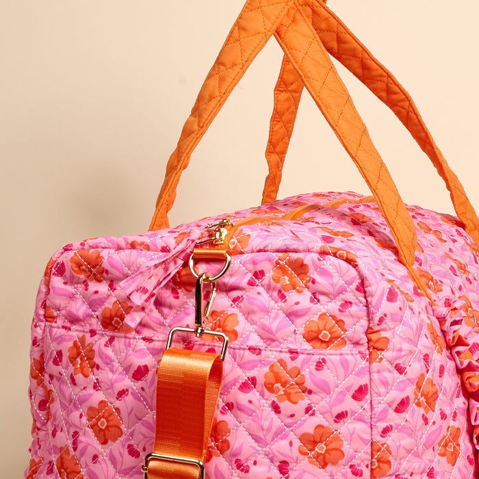 A close-up of The Darling Effect's Poppin' Petals Hot Pink On-the-Go Quilted Weekender Bag, featuring pink and orange floral design, orange handles, and an adjustable shoulder strap with gold clasp on a beige background.