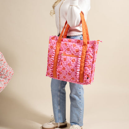A woman in a white sweater and blue jeans carries The Darling Effect's Poppin' Petals Hot Pink The Everyday Quilted Tote Bag, featuring orange handles, ruffle details, an exterior zip pocket, and styled casually with sneakers.