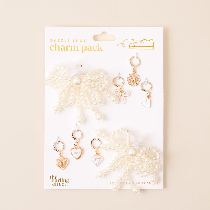 The Pretty in Pearl Dazzle Shoe Charm Pack by the-darling-effect includes two large pearl bows and six gold and white sneaker charms—hearts, diamond, bow, flower, circle—on a labeled white card. Ideal for footwear accessories lovers.