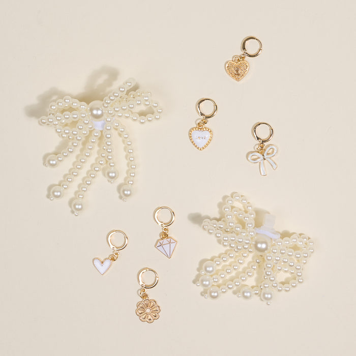 Two pearl bow hair clips and seven gold and white heart, bow, flower, and diamond-shaped charms from the-darling-effect’s Pretty in Pearl Dazzle Shoe Charm Pack are arranged on a light beige surface.