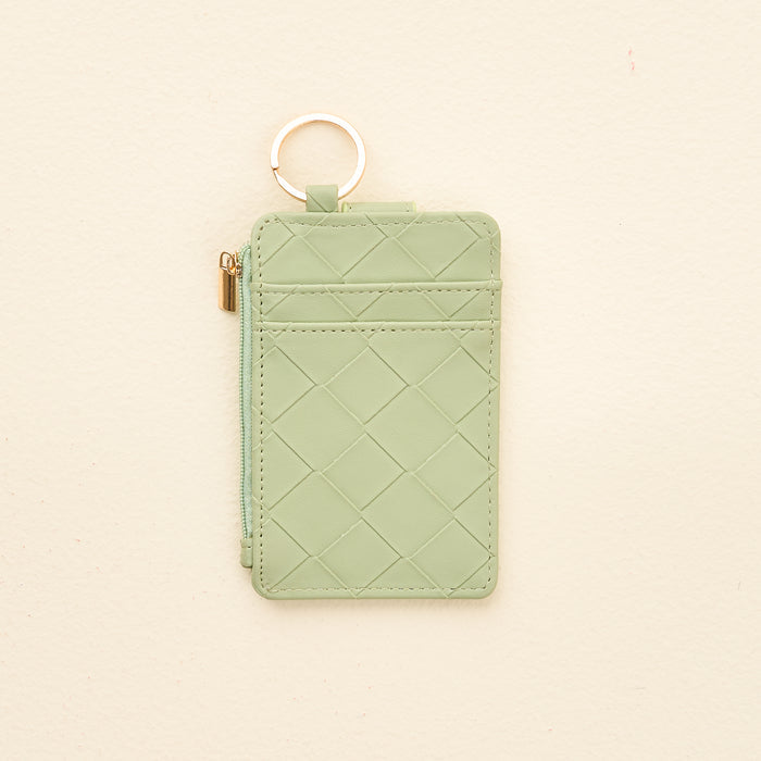 The Darling Effect’s Sage Woven Keychain Wallet features a quilted light green design, gold keyring, and zipper—all in a compact accessory—shown on a neutral beige background.