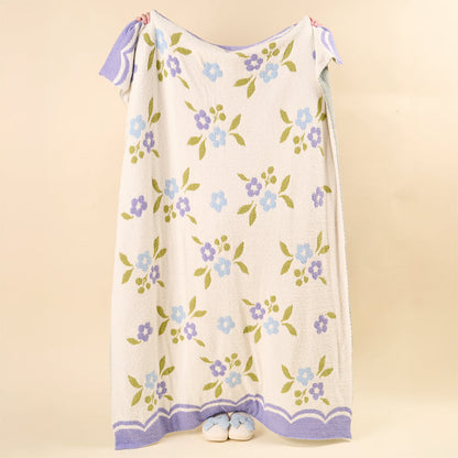 A person is concealed behind a Scalloped Floral Periwinkle Cozy Dreams Luxe Blanket by the-darling-effect, with only their feet in white slippers peeking out at the bottom against a solid beige background.