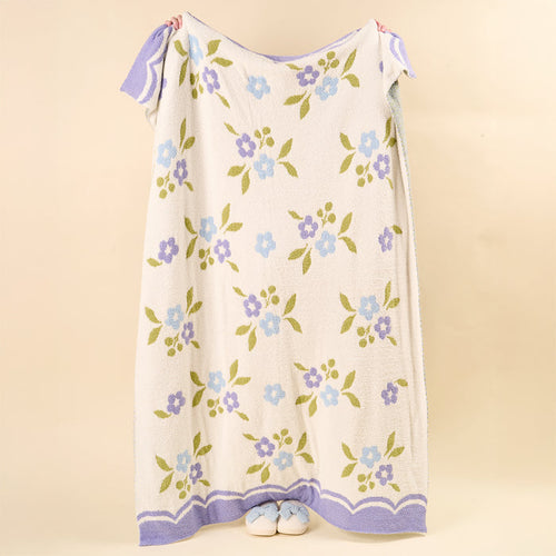 A person is concealed behind a Scalloped Floral Periwinkle Cozy Dreams Luxe Blanket by the-darling-effect, with only their feet in white slippers peeking out at the bottom against a solid beige background.