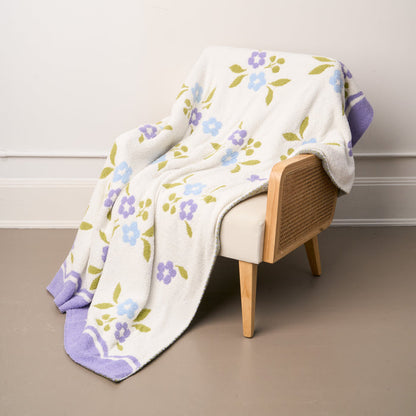 A white armchair with cane sides rests on a taupe floor, draped with the-darling-effect’s Scalloped Floral Periwinkle Cozy Dreams Luxe Blanket—an ultra-soft microfiber blanket adorned with purple and blue flowers and finished with a lavender scalloped trim.