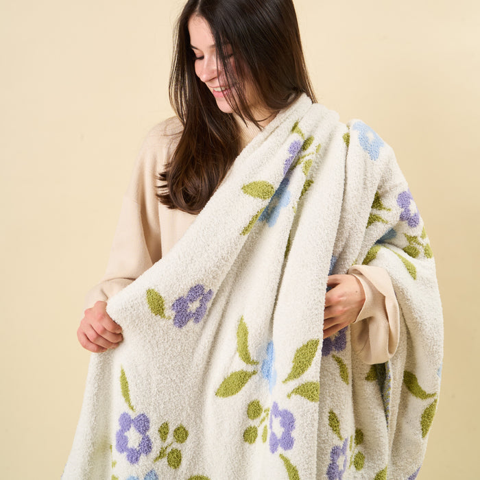 A woman with long dark hair smiles while wrapping herself in the Scalloped Floral Periwinkle Cozy Dreams Luxe Blanket from the-darling-effect, an ultra-soft, fluffy white blanket adorned with purple and blue flowers and green leaves. The background is a plain light beige color.