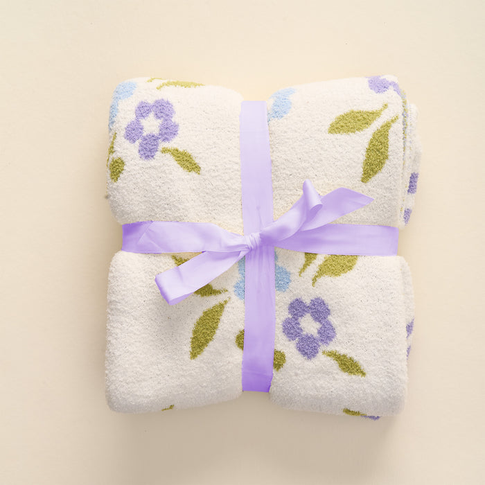 A white towel adorned with purple and green floral patterns is neatly folded and tied with a light purple ribbon, set against a light background—perfect for pairing with your the-darling-effect Scalloped Floral Periwinkle Cozy Dreams Luxe Blanket for an added touch of luxury.