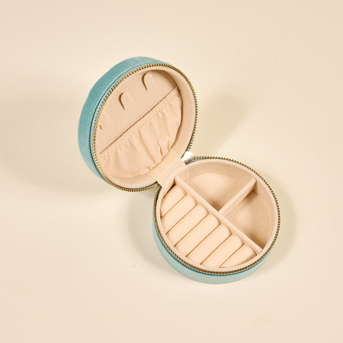 The Darling Effect Seashell Round Velvet Jewelry Case in light blue features a zip closure and opens to a beige interior with ring slots, small compartments, and a notched pocket for organized jewelry storage.