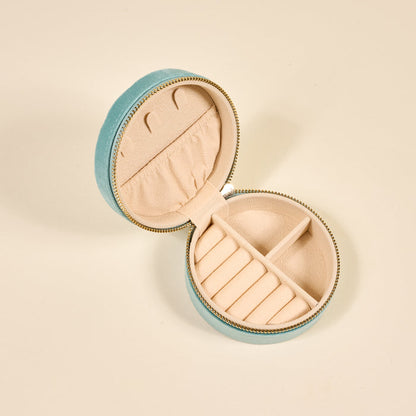 The Darling Effect Seashell Round Velvet Jewelry Case in light blue features a zip closure and opens to a beige interior with ring slots, small compartments, and a notched pocket for organized jewelry storage.