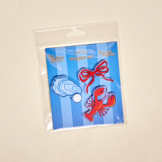 The Darling Effect's Seaside Haven Little Magnet Set features three acrylic magnets—blue oyster, red lobster, and red bow—on a blue and white striped card in a clear bag, perfect for decorating your fridge.