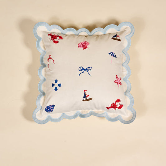 The Darling Effect Seaside Haven Scalloped Square Throw Pillow features light blue scalloped edges and nautical embroidery—lobster, sailboat, shell, anchor, bow, life preserver, umbrella—on a cream background for a cozy touch.