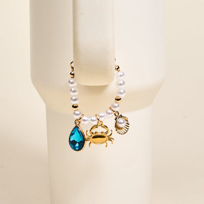 The Darling Effect’s Seaside Sparkle Tumbler Charm features pearls, gold beads, and three ocean charms—blue teardrop crystal, gold crab, and scallop shell with pearl—perfect for personalizing your favorite drinkware. Shown on a white mannequin wrist.