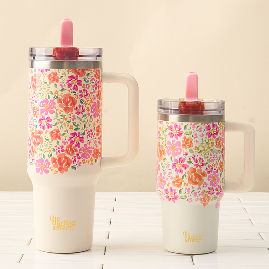 The Secret Garden Pink 40 oz Lifestyle Flip Straw Tumbler by the-darling-effect features bright pink, orange, and peach floral patterns on cream, has a handle and leak-resistant lid, and displays the darling effect logo at the bottom.