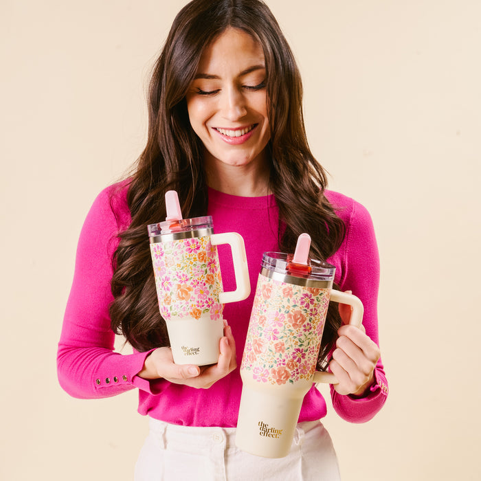 A woman in a bright pink top smiles while holding two the-darling-effect Secret Garden Pink 40 oz Lifestyle Flip Straw Tumblers, featuring handles, straws, and a stylish leak-resistant design against a beige background.
