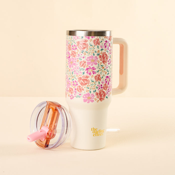 The Secret Garden Pink 40 oz Lifestyle Flip Straw Tumbler by the-darling-effect, featuring a colorful floral pattern, a leak-resistant clear lid with pink straw, and yellow branding, sits on a light surface with its handle visible.