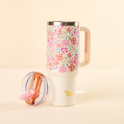 The Secret Garden Pink 40 oz Lifestyle Flip Straw Tumbler by the-darling-effect, featuring a colorful floral pattern, a leak-resistant clear lid with pink straw, and yellow branding, sits on a light surface with its handle visible.