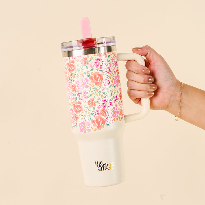 A hand holds the-darling-effect Secret Garden Pink 40 oz Lifestyle Flip Straw Tumbler with a floral pattern, handle, clear leak-resistant lid, red flip straw, and logo on the cream-colored bottom section.