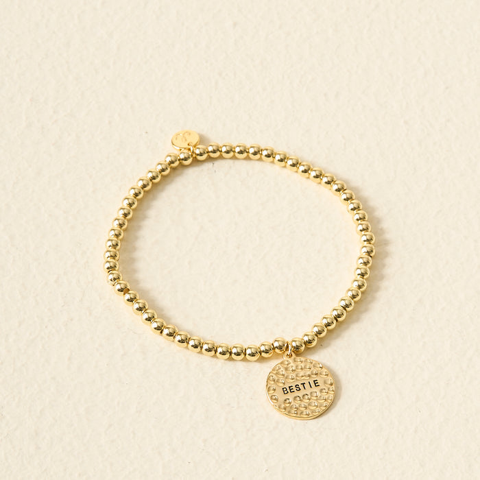 The Darling Effect’s Sentiment Beaded Charm Bracelet features gold beads and a round BESTIE-engraved charm, making it a thoughtful gift, beautifully showcased on a light textured background.