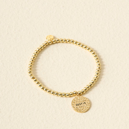 The Darling Effect’s Sentiment Beaded Charm Bracelet features gold beads and a round BESTIE-engraved charm, making it a thoughtful gift, beautifully showcased on a light textured background.