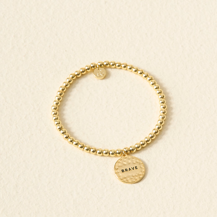 The Darling Effect Sentiment Beaded Charm Bracelet features gold beads and a round BRAVE-engraved charm, displayed on a light textured background—a meaningful gift to inspire courage.