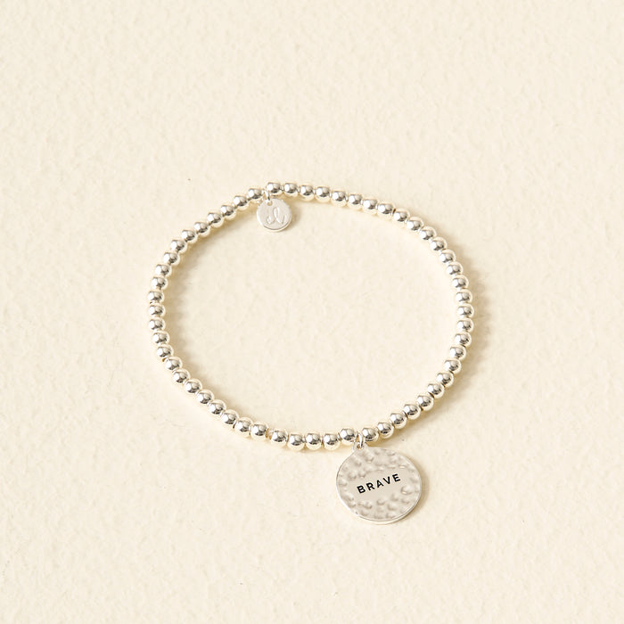 The Darling Effect Sentiment Beaded Charm Bracelet features silver beads and a round, hammered BRAVE charm—an ideal sentimental gift, beautifully displayed on a light, textured surface.
