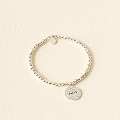 The Darling Effect Sentiment Beaded Charm Bracelet features silver beads and a round, hammered BRAVE charm—an ideal sentimental gift, beautifully displayed on a light, textured surface.